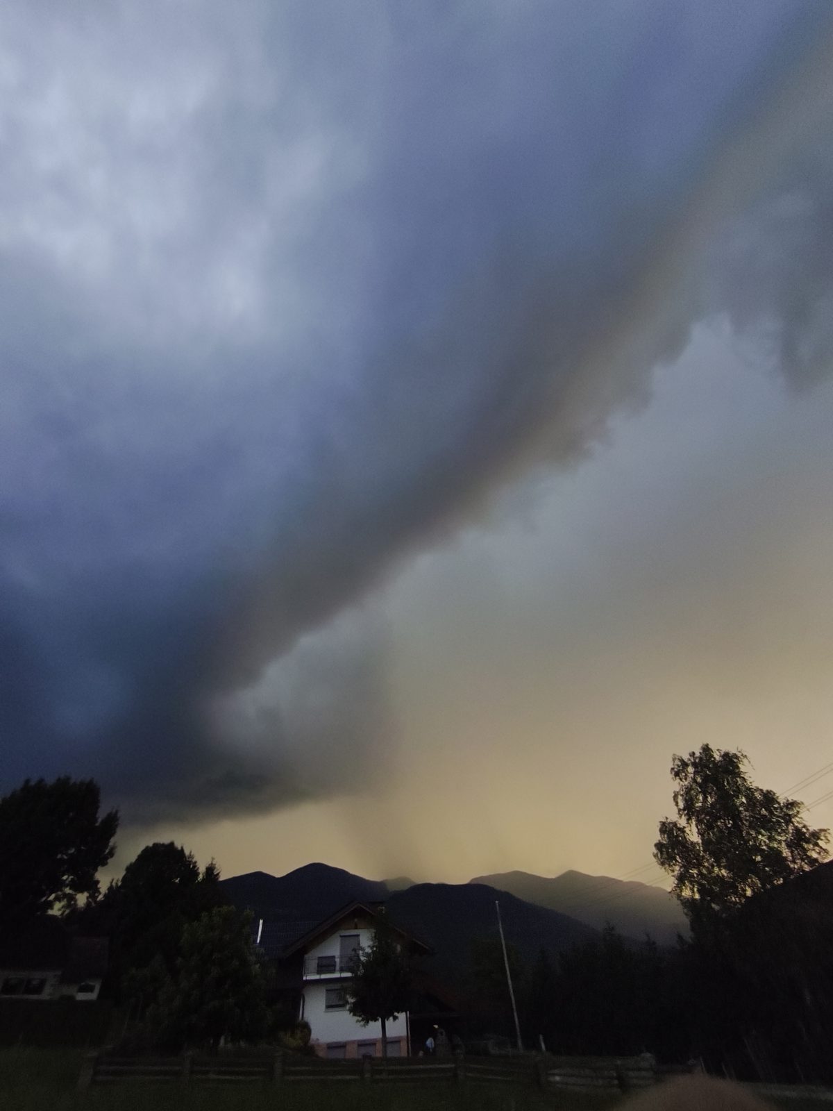 Padavinski curek (rain shaft) pod nevihtnim oblakom nad pogorjem, obarvan z večerno svetlobo