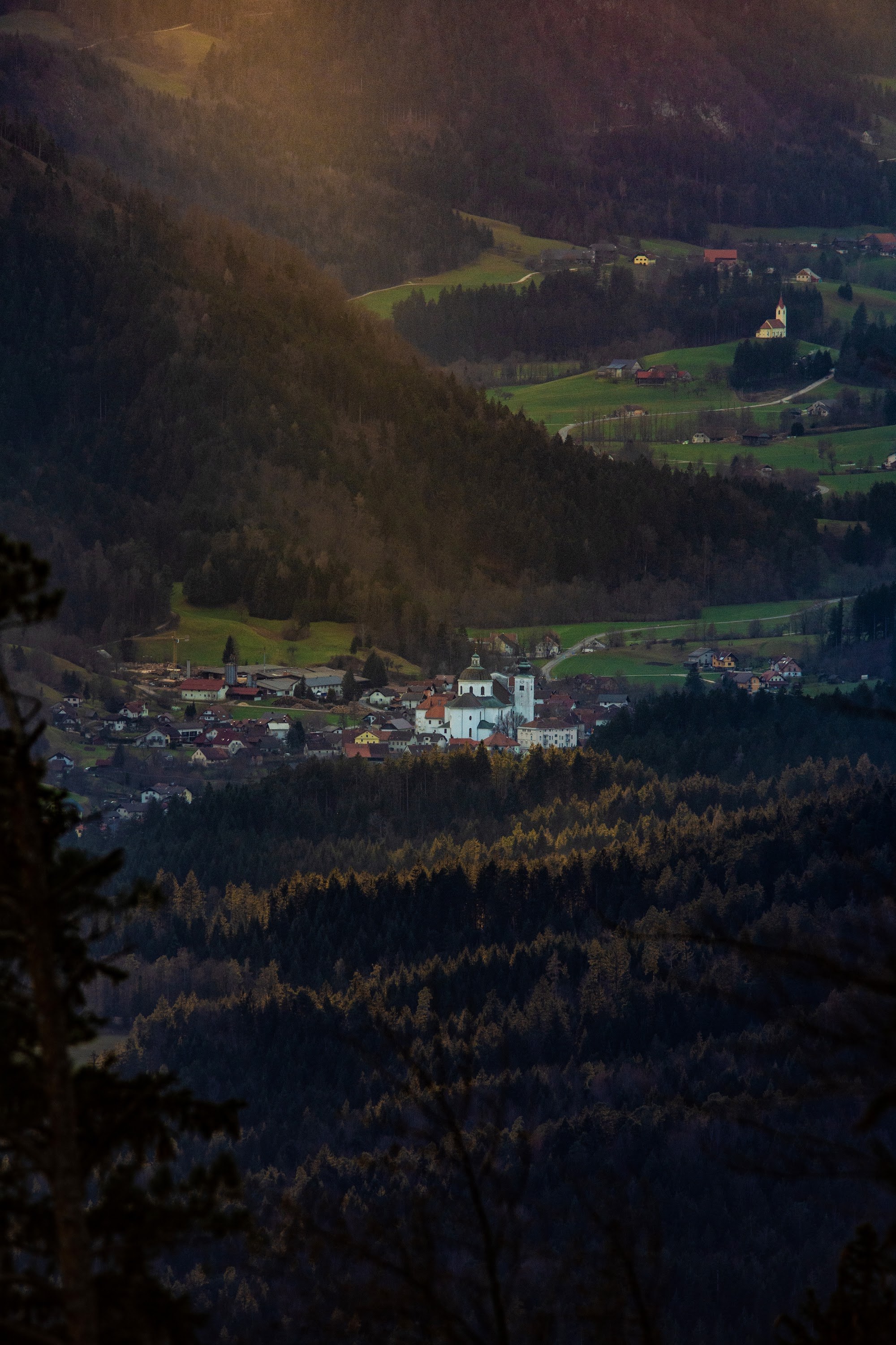 Večerna svetloba nad vasjo v Zgornji Savinjski dolini, obkroženo z gozdnatimi pobočji.