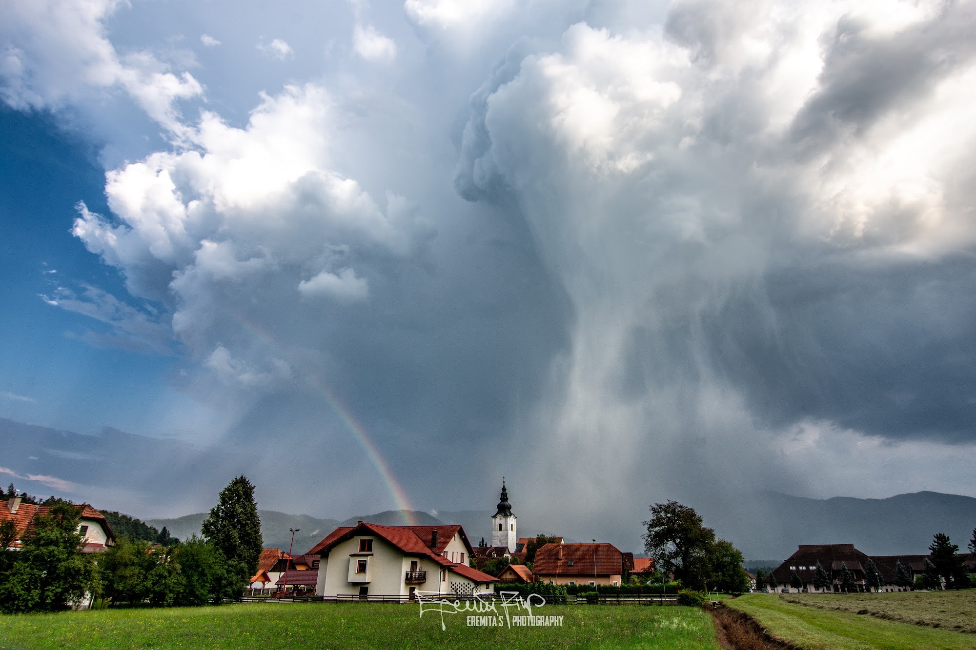 Osamljena poletna nevihta z izrazitim padavinskim curkom in mavrico nad vasjo.